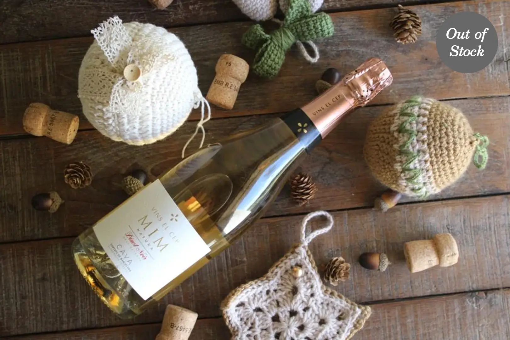 ```json { "alt": "A bottle of sparkling wine on a wooden table with knitted decorations, corks, and pinecones, marked out of stock.", "title": "Sparkling Wine with Knitted Decor and Corks on a Table" } ```